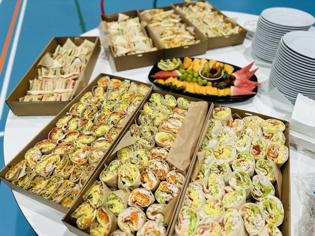 7 larges boxes of sandwiches and wraps on a table with a fruit platter in the middle and stacks of plates